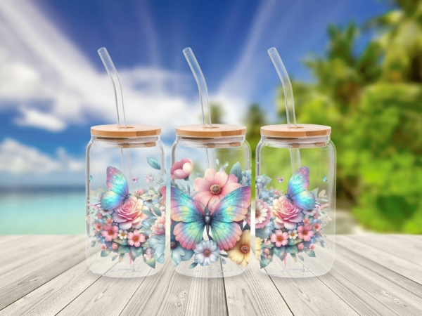 Drei 16 oz Glasbecher mit Bambusdeckel und Glastrinkhalm, verziert mit lebendigen Blumenmotiven und schillernden Schmetterlingen in Pastelltönen. Die Becher stehen auf einem Holztisch vor einem tropischen Strandhintergrund, was eine fröhliche und naturver